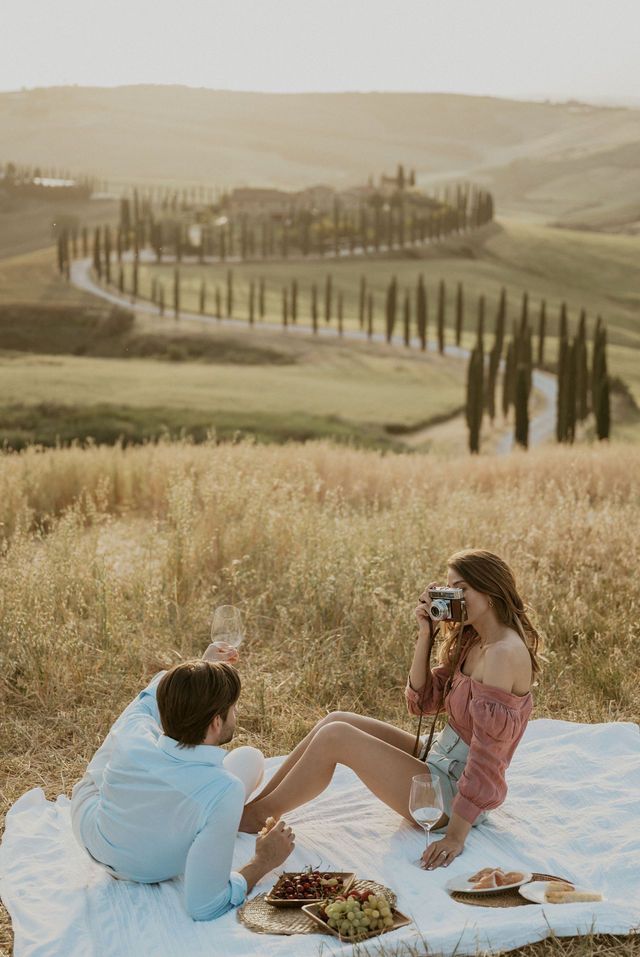 Picnic scene in Tuscan countryside with person taking photos against cypress tree backdrop.