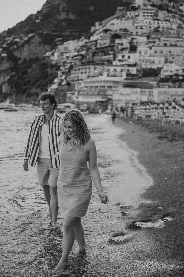 Couple walking along beach with Positano's cascading buildings in background.