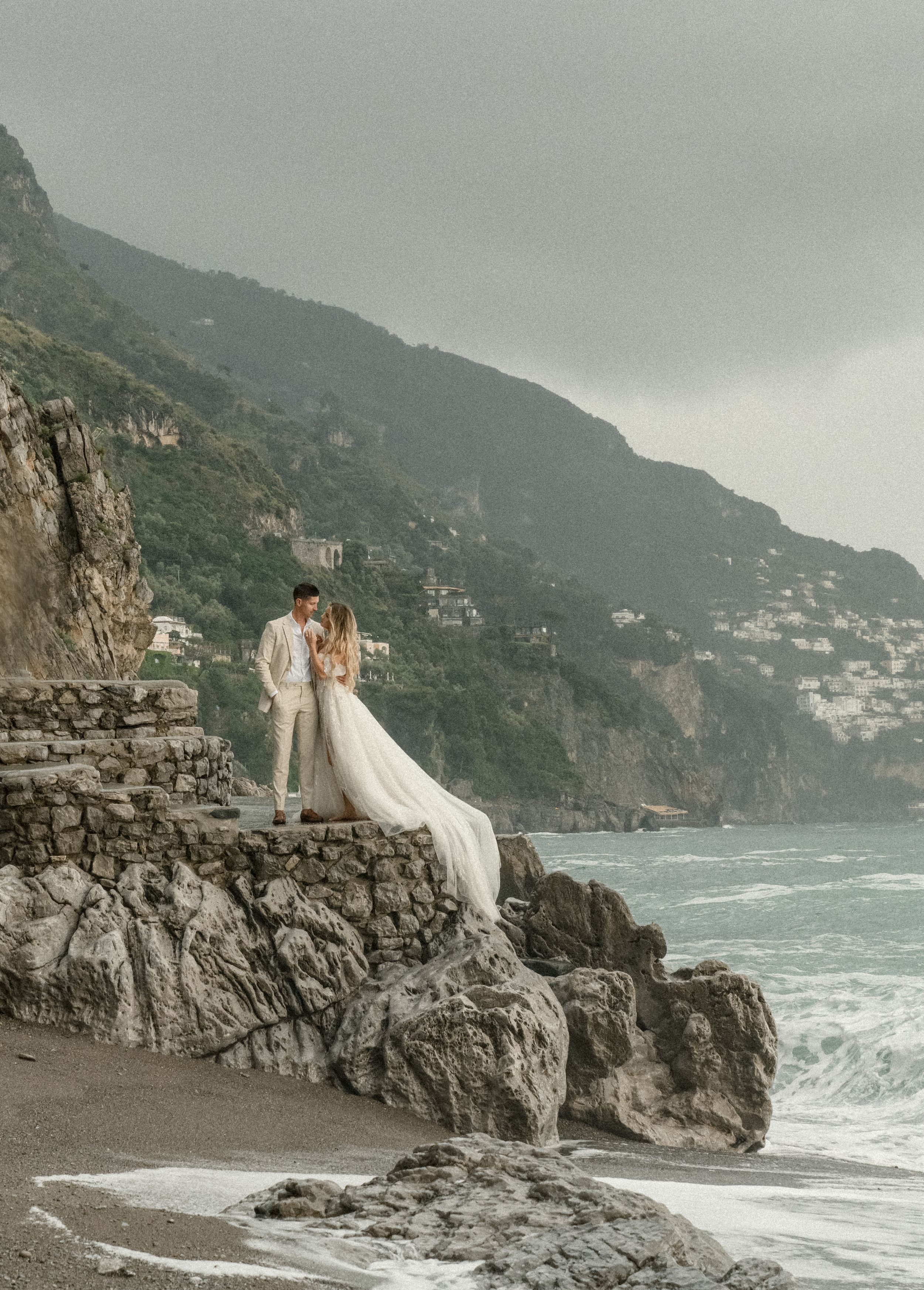 A couple poses on rocky coastal cliffs with dramatic mountainous coastline in the background.
