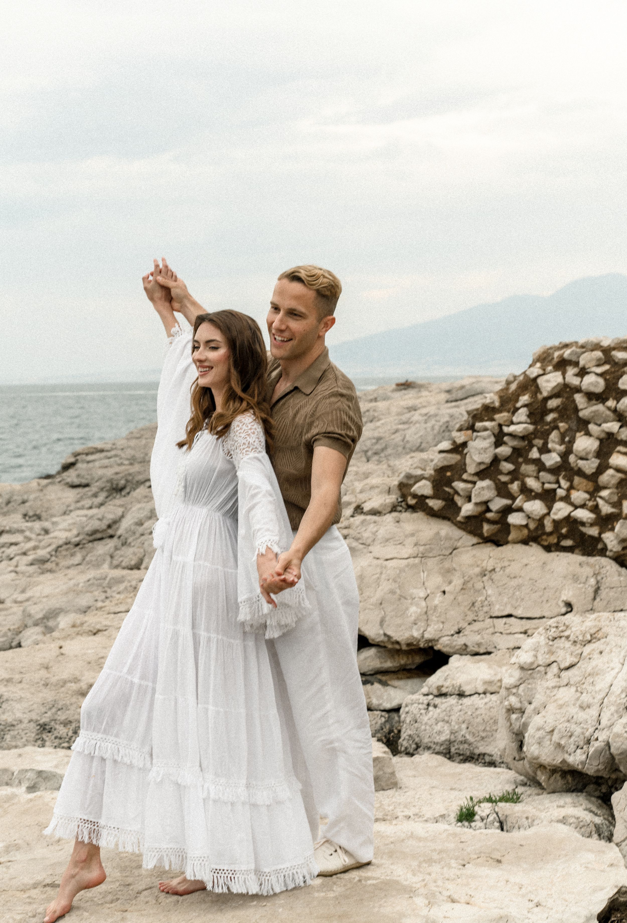 Romantic couple poses on rocky coastal terrain with white dress and casual attire against ocean backdrop.
