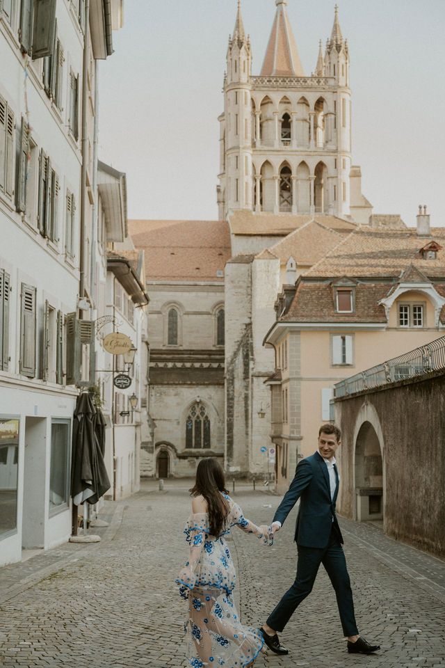 Couple walking hand in hand through cobblestone street with Gothic cathedral tower in background.