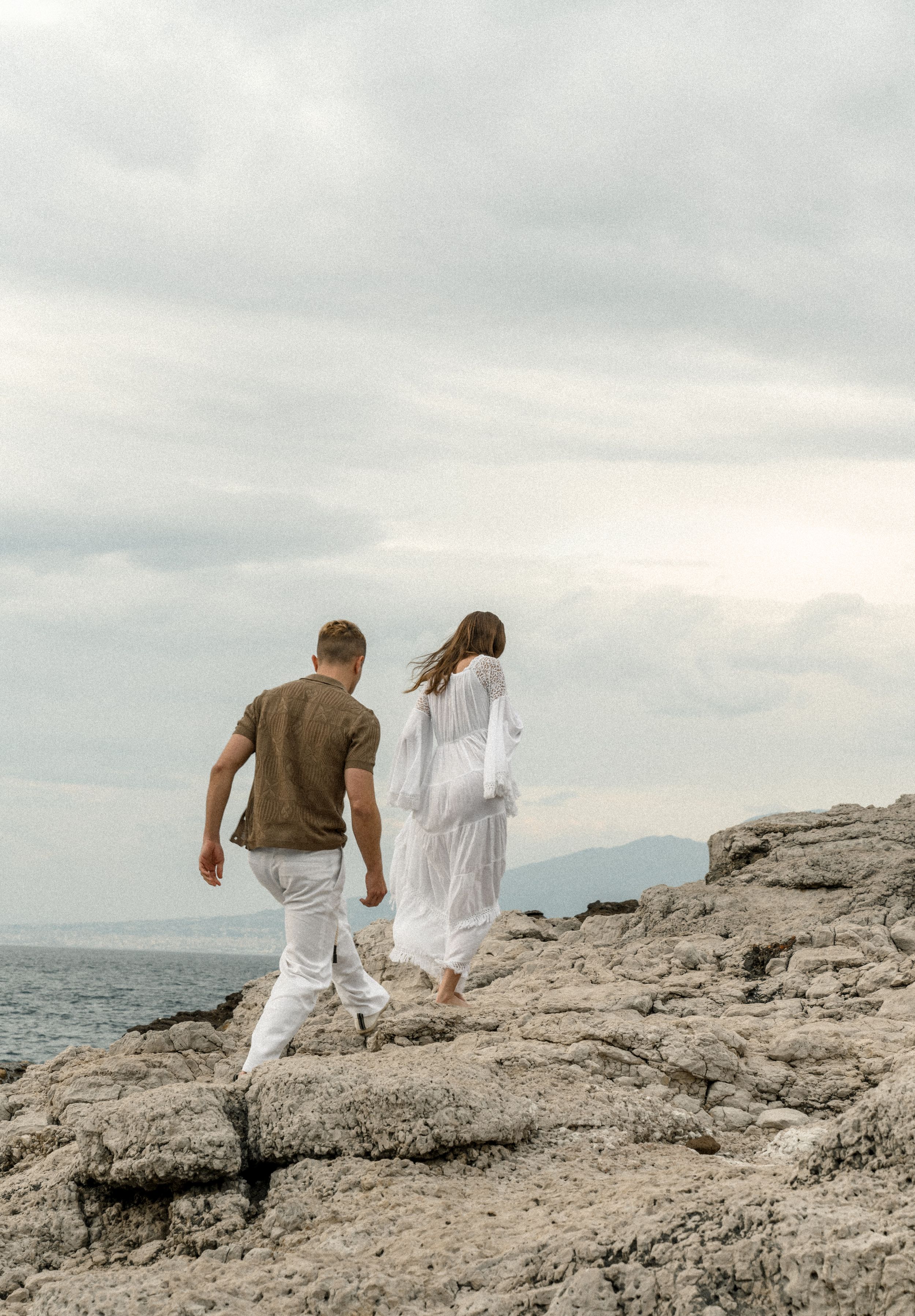 Couple walks along rocky coastline wearing white and brown outfits against cloudy sky.