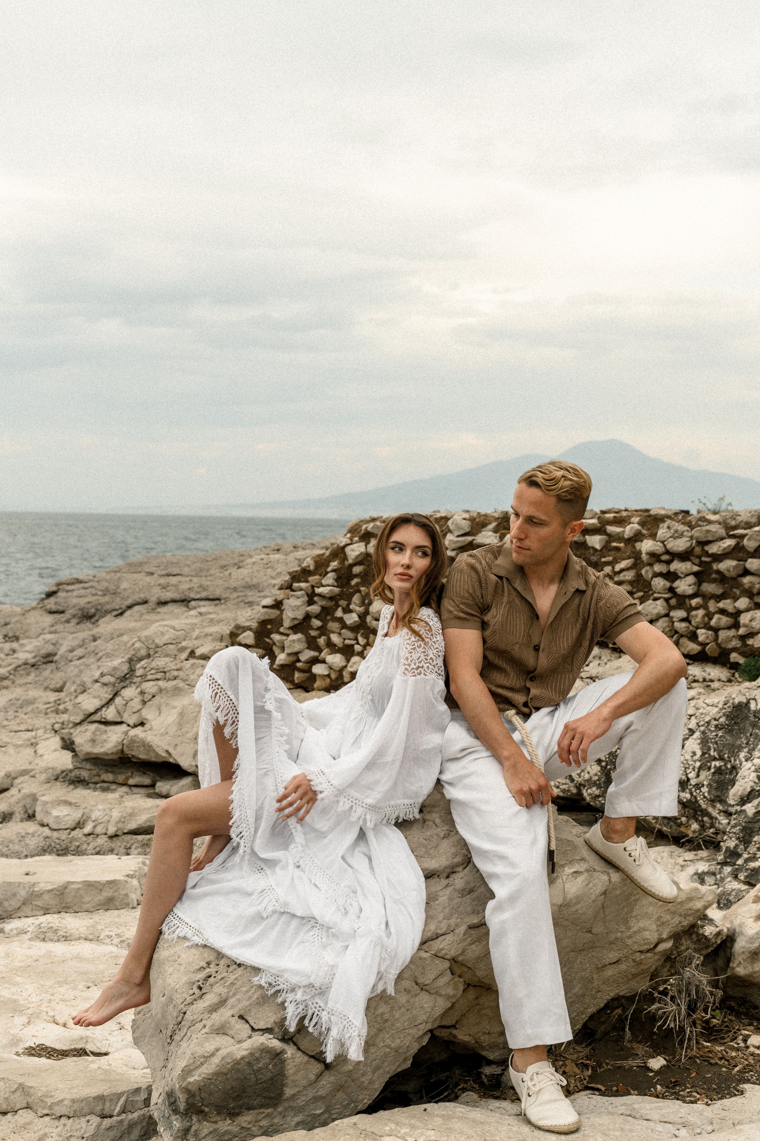 Couple relaxes on rocky coastline wearing white dress and casual outfit with mountains in background.