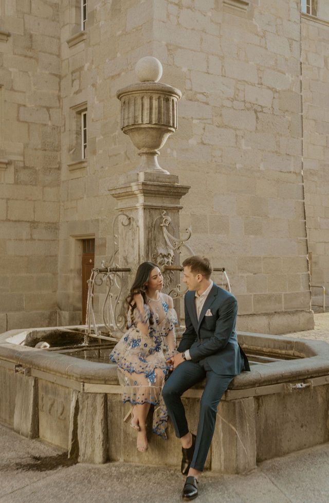 Romantic couple sitting on historic stone fountain in European courtyard during engagement shoot.