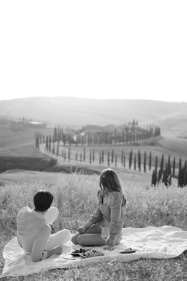 Couple having romantic picnic overlooking rolling Tuscan hills with cypress trees in black and white.