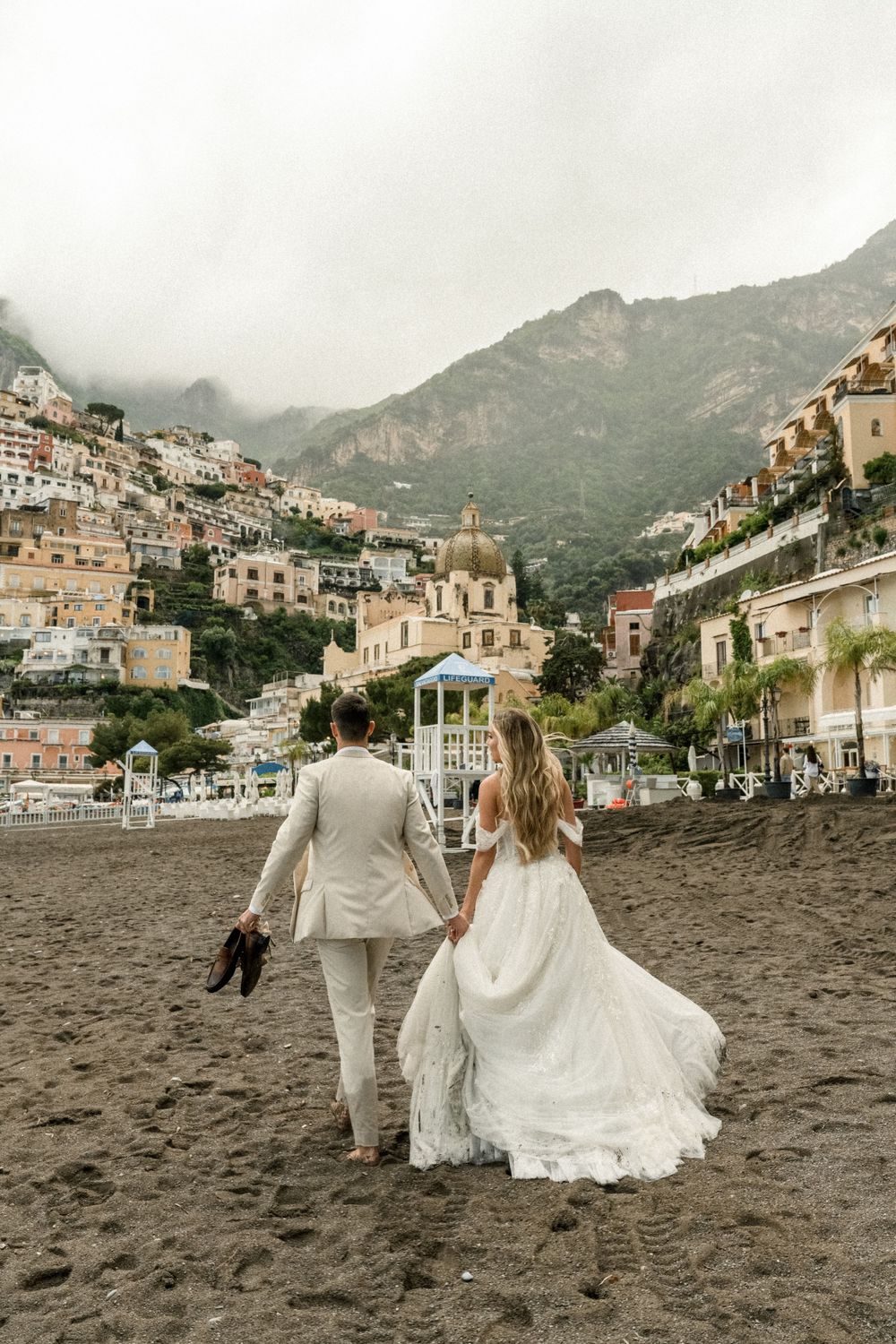 A couple walks on a black sand beach in front of the stunning cliffside town of Positano, Italy.