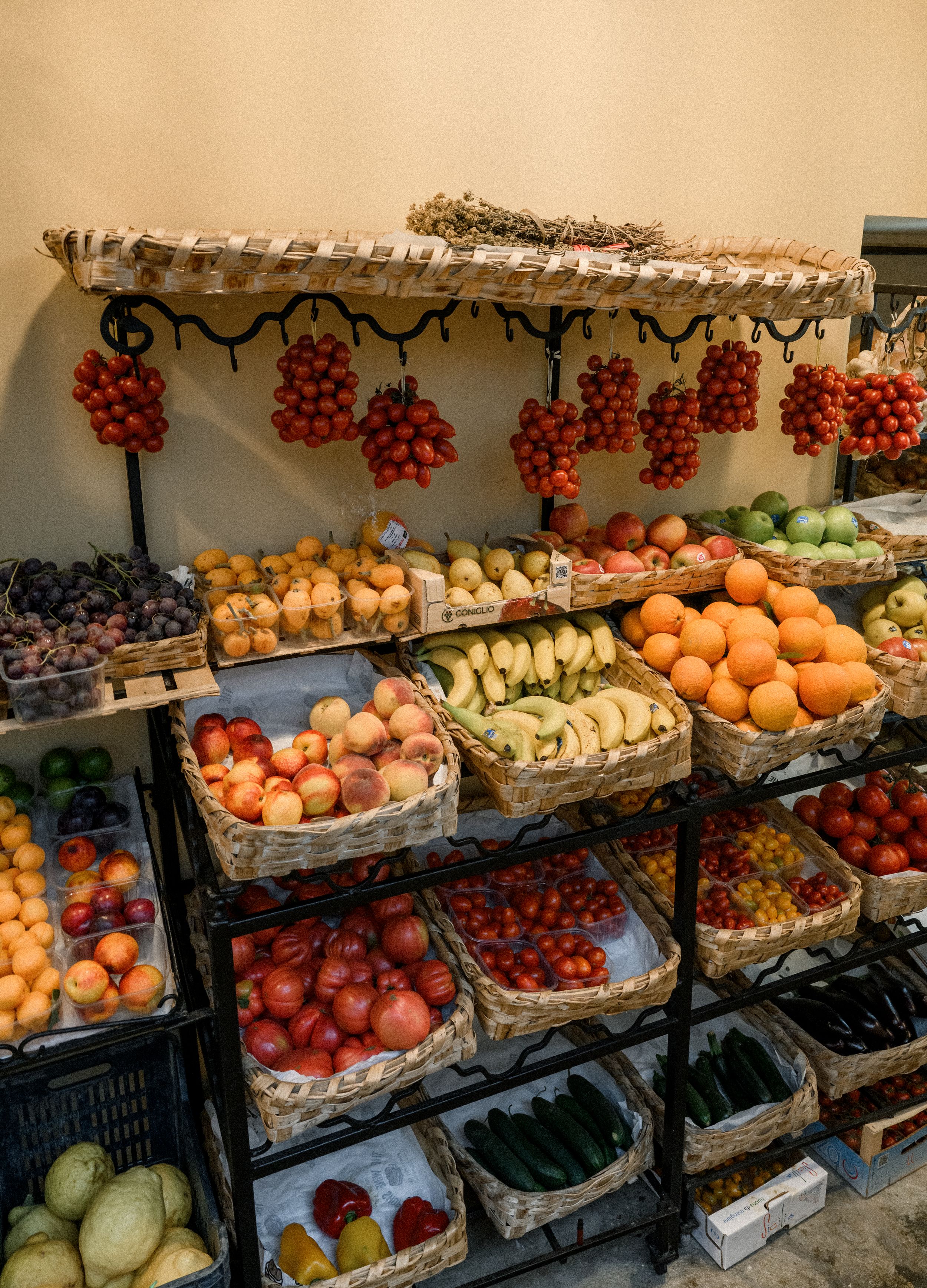 Colorful fresh produce display at a local market with hanging tomatoes and stacked fruits in baskets.