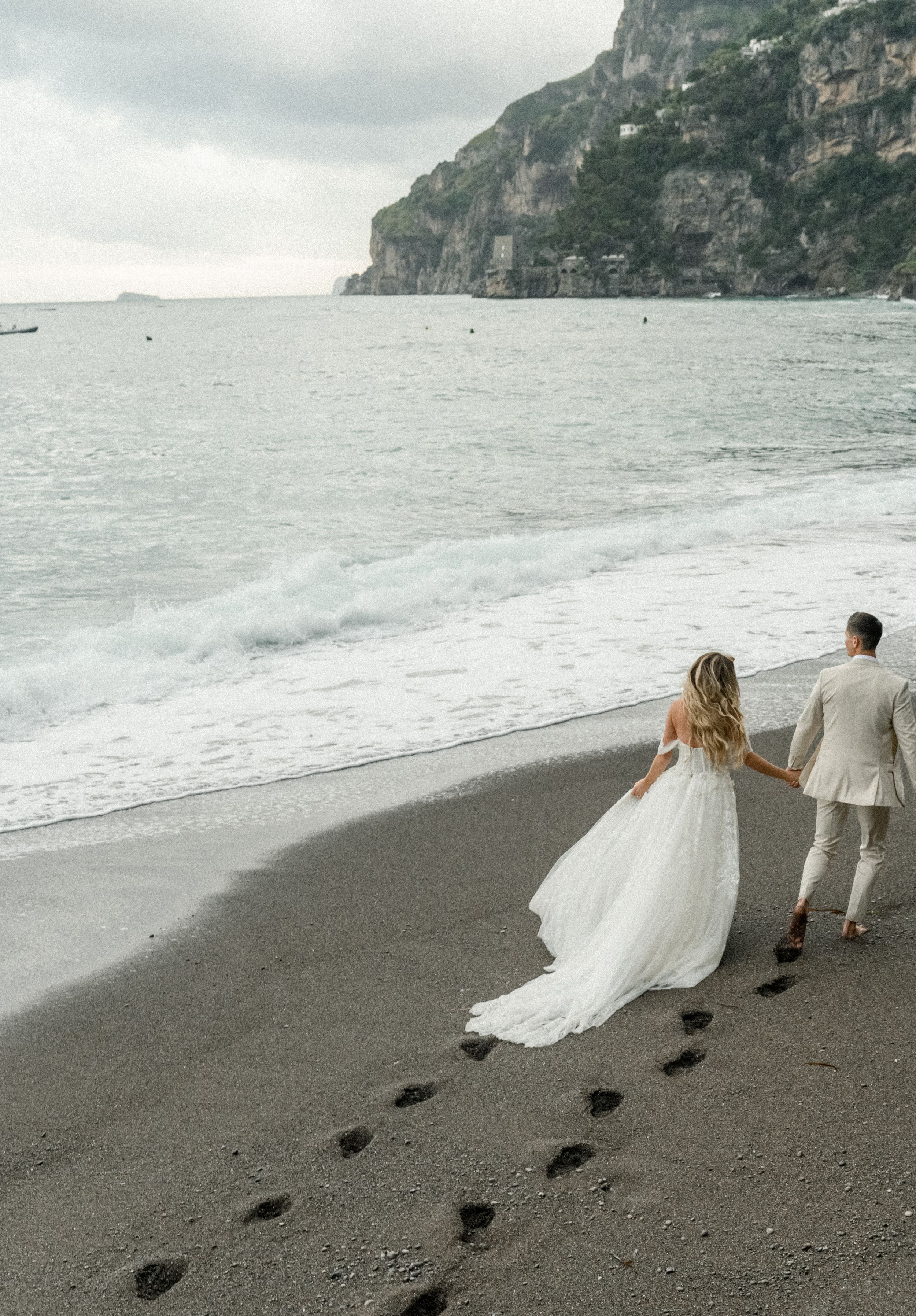 Couple walking hand in hand leaving footprints on dark sandy beach with cliffs in background.