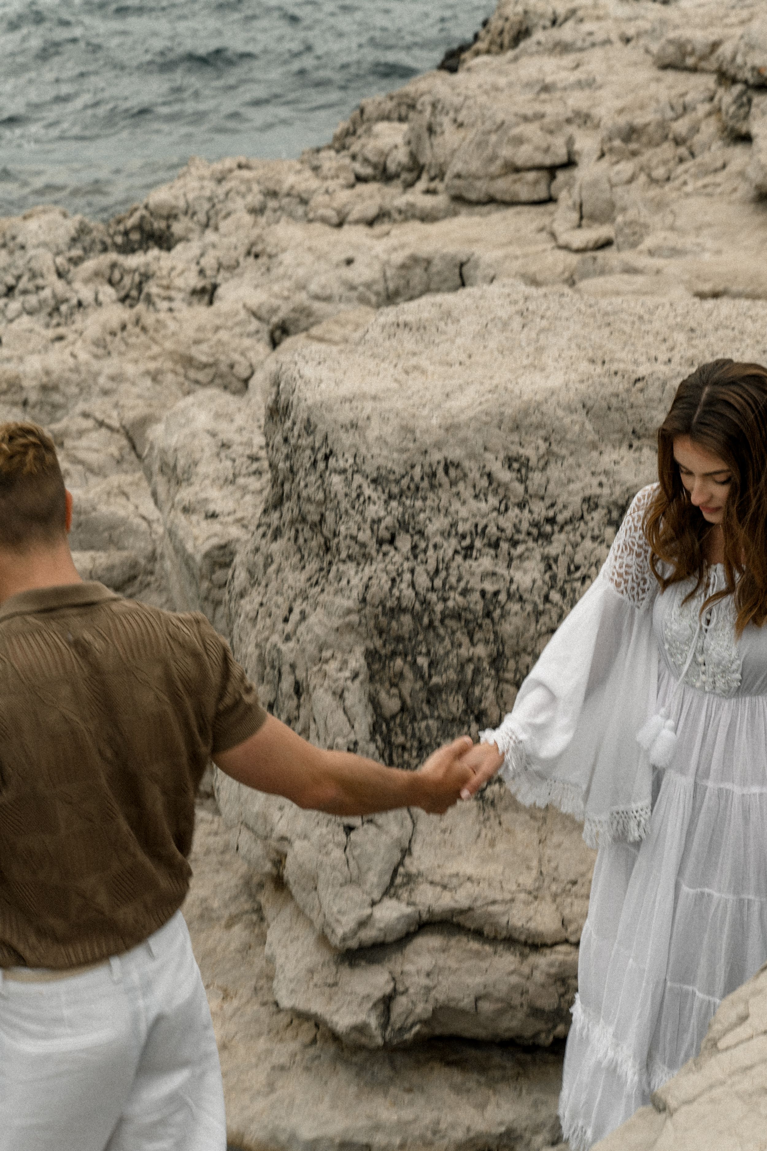 Couple holding hands on rocky coastal terrain, white flowing dress contrasting against natural landscape.