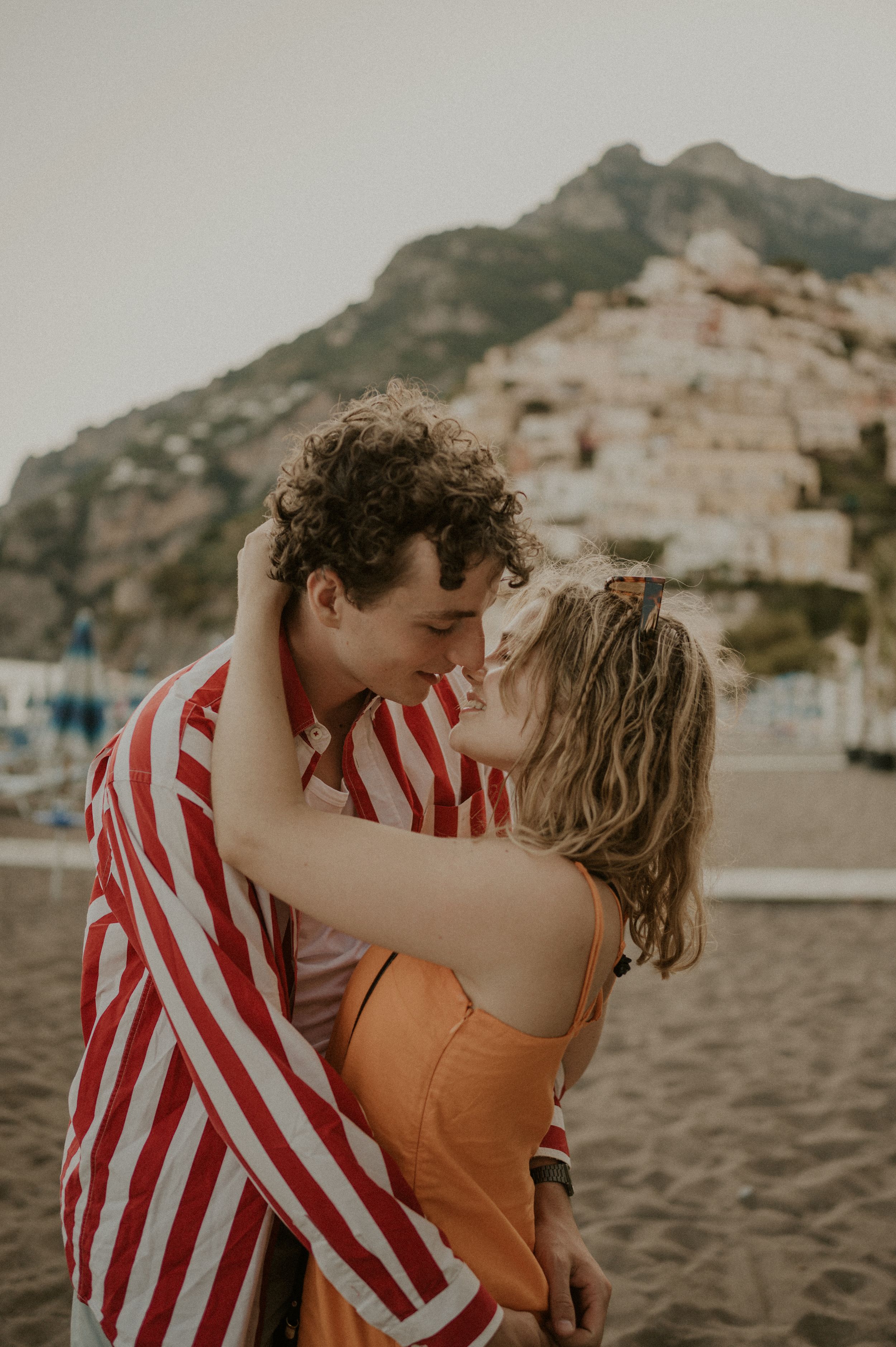 A romantic moment captured on a beach with Mediterranean coastal buildings rising up the hillside.