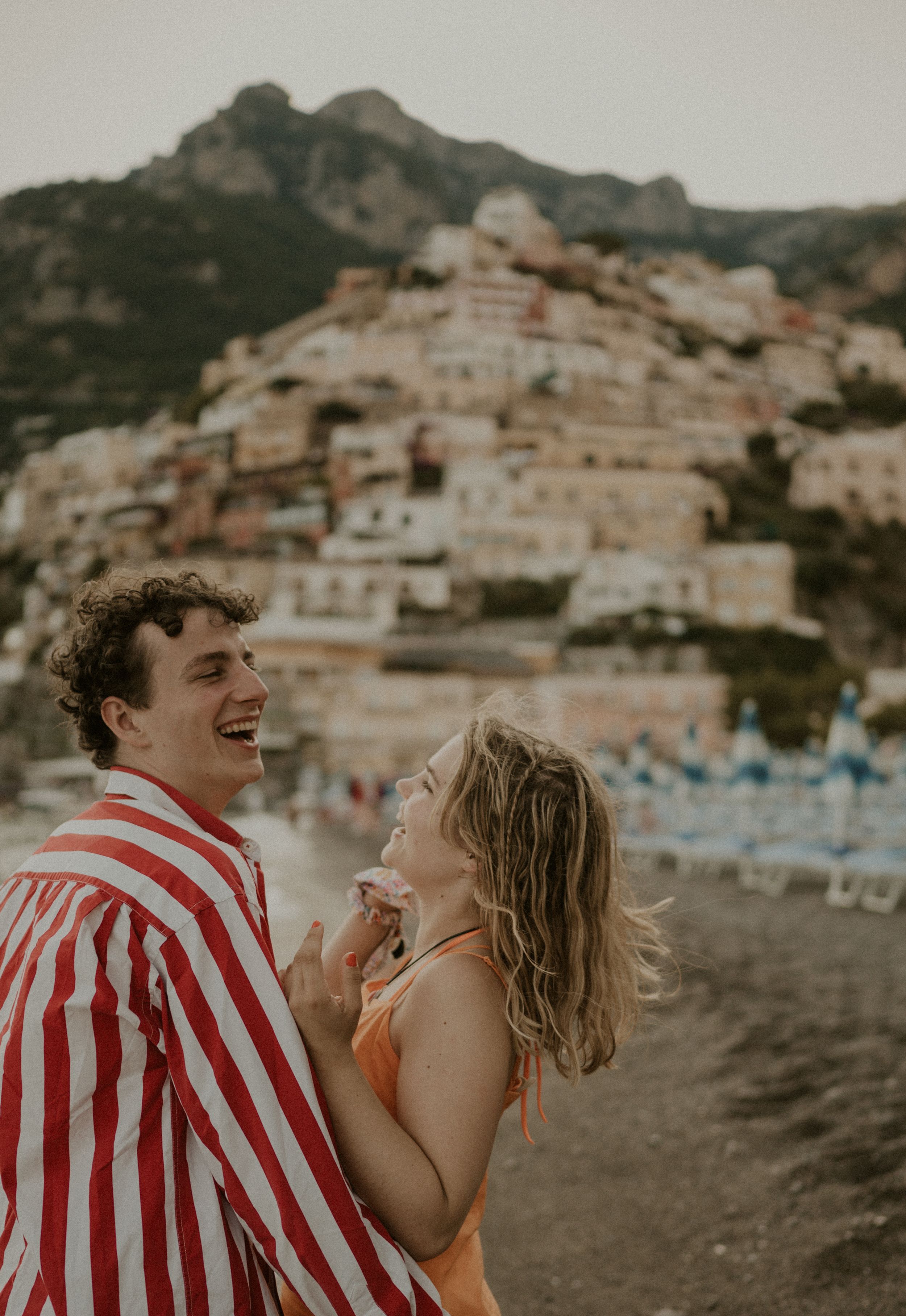A couple laughs together on a beach with the colorful hillside town of Positano, Italy in the background.