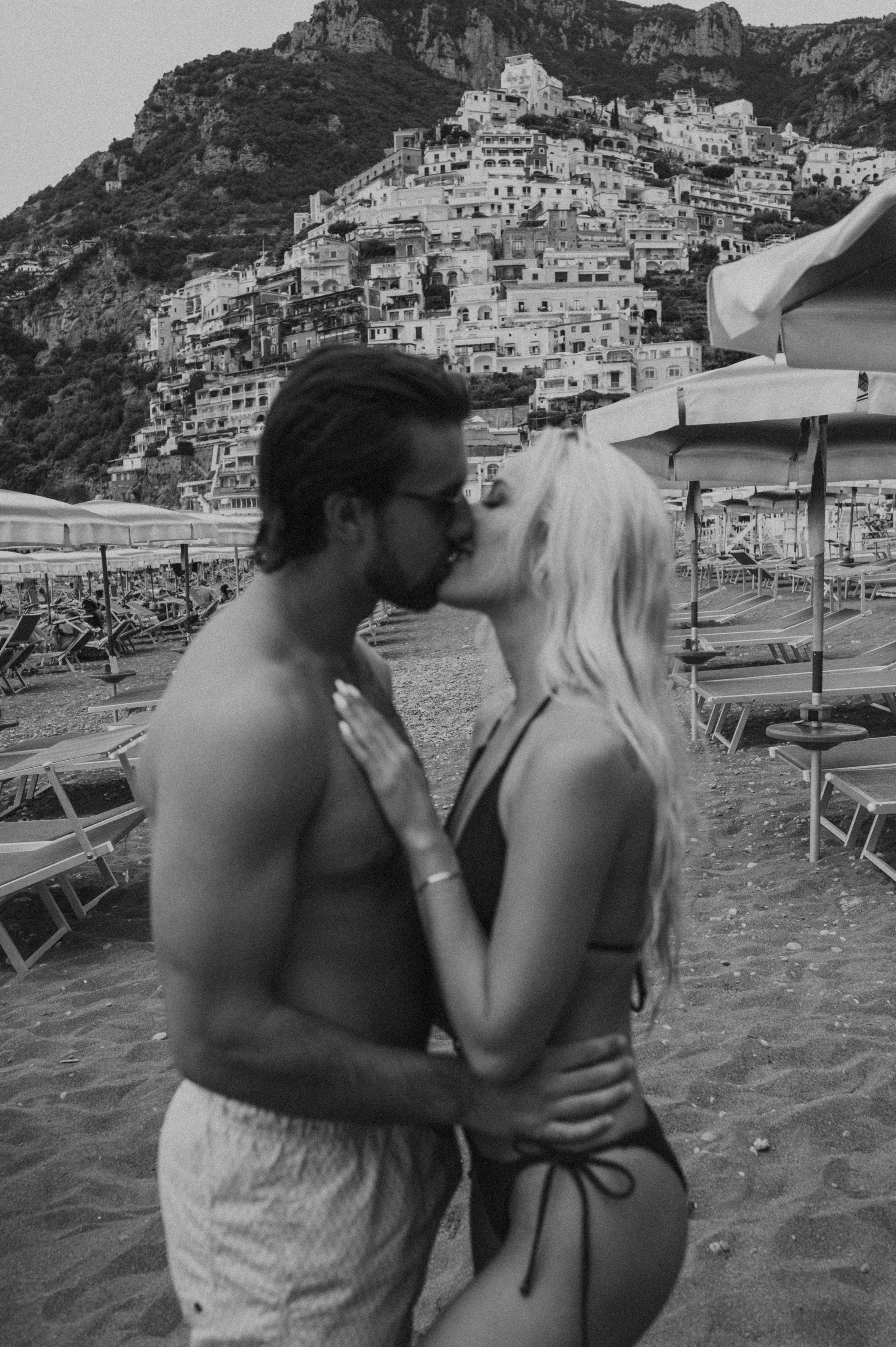 A romantic scene at a beach in Positano with the iconic hillside town buildings cascading down the cliffside in black and white.