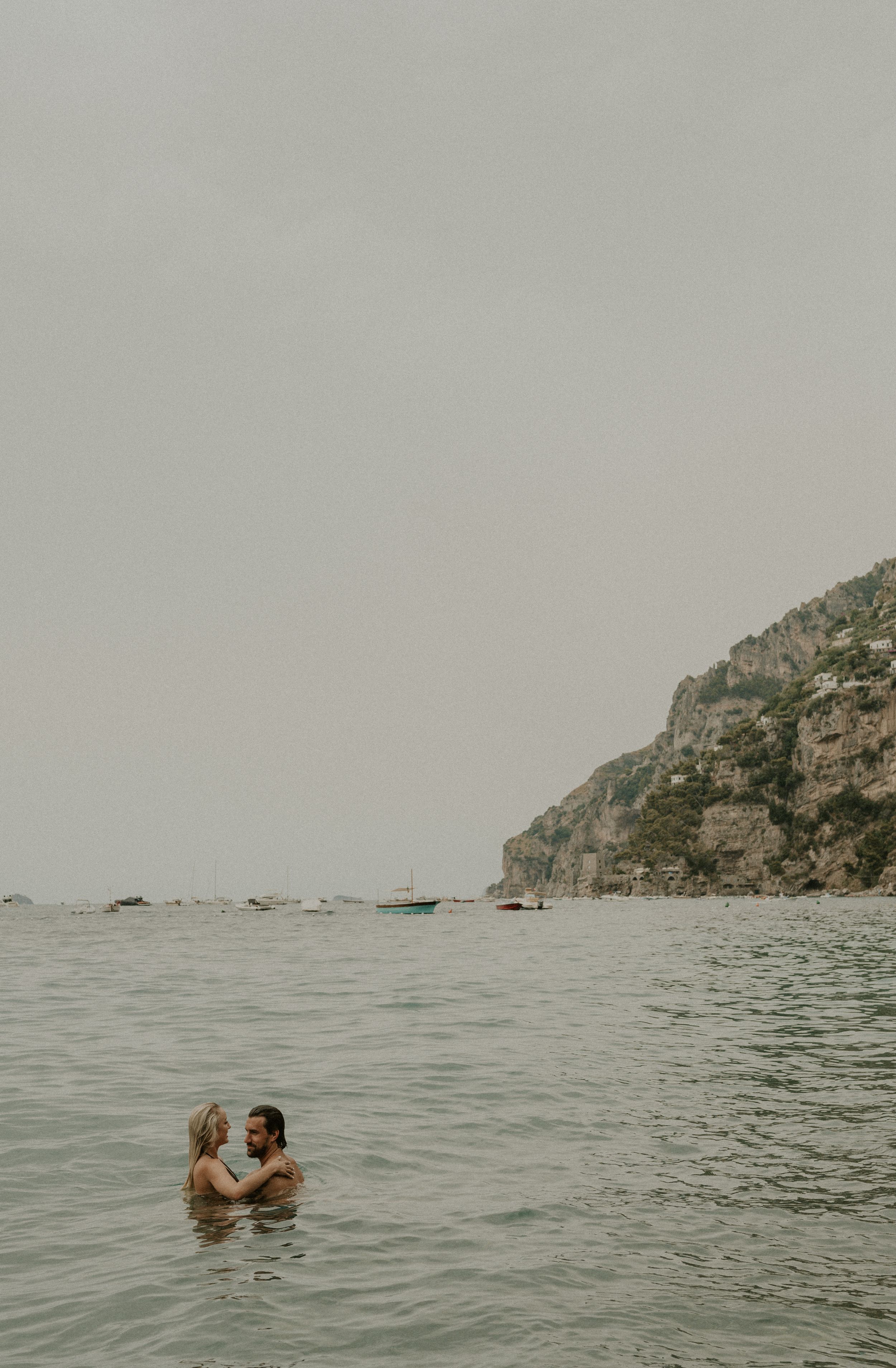 Couple embracing in Mediterranean waters with coastal cliffs in background.