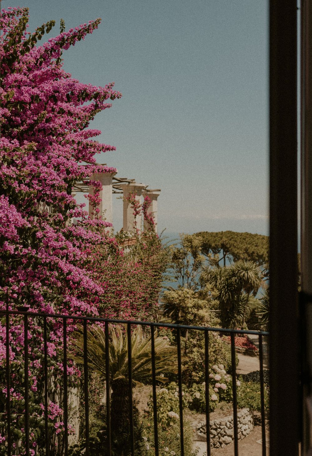 Pink bougainvillea framing ancient stone columns against a Mediterranean coastline.