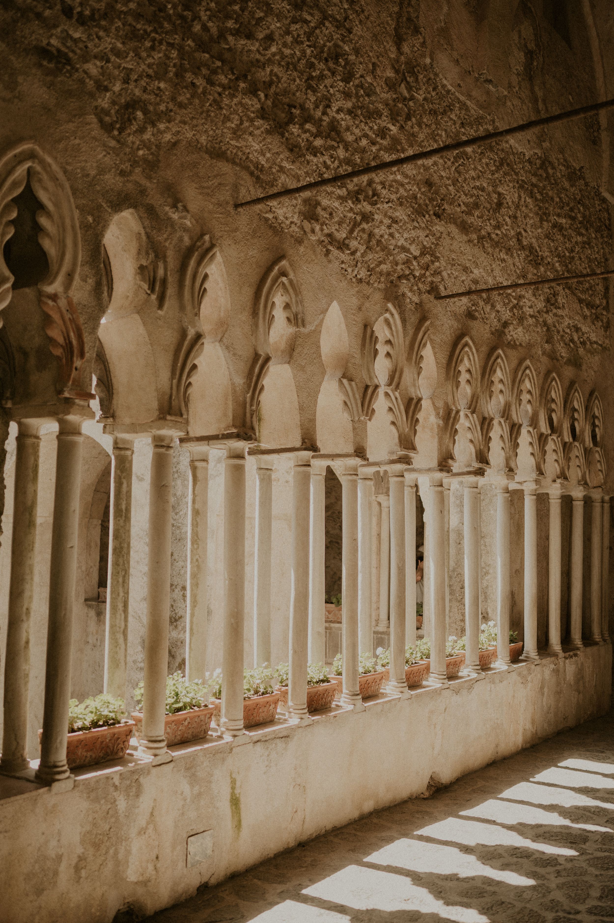 Historic stone colonnade with arched windows and potted plants in sepia tones.