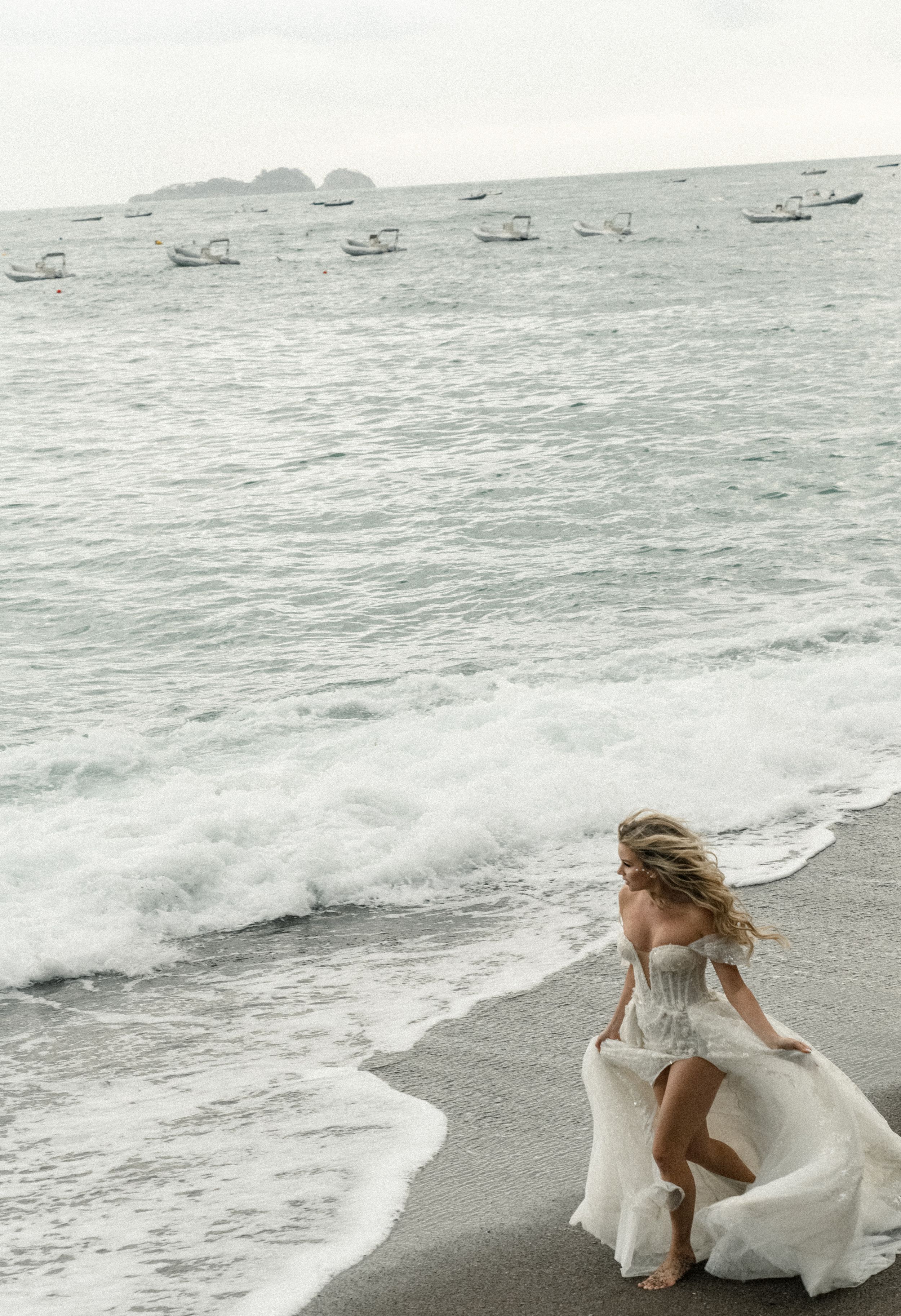A white dress flows in the ocean breeze as waves crash on a rocky beach with distant boats on the horizon.