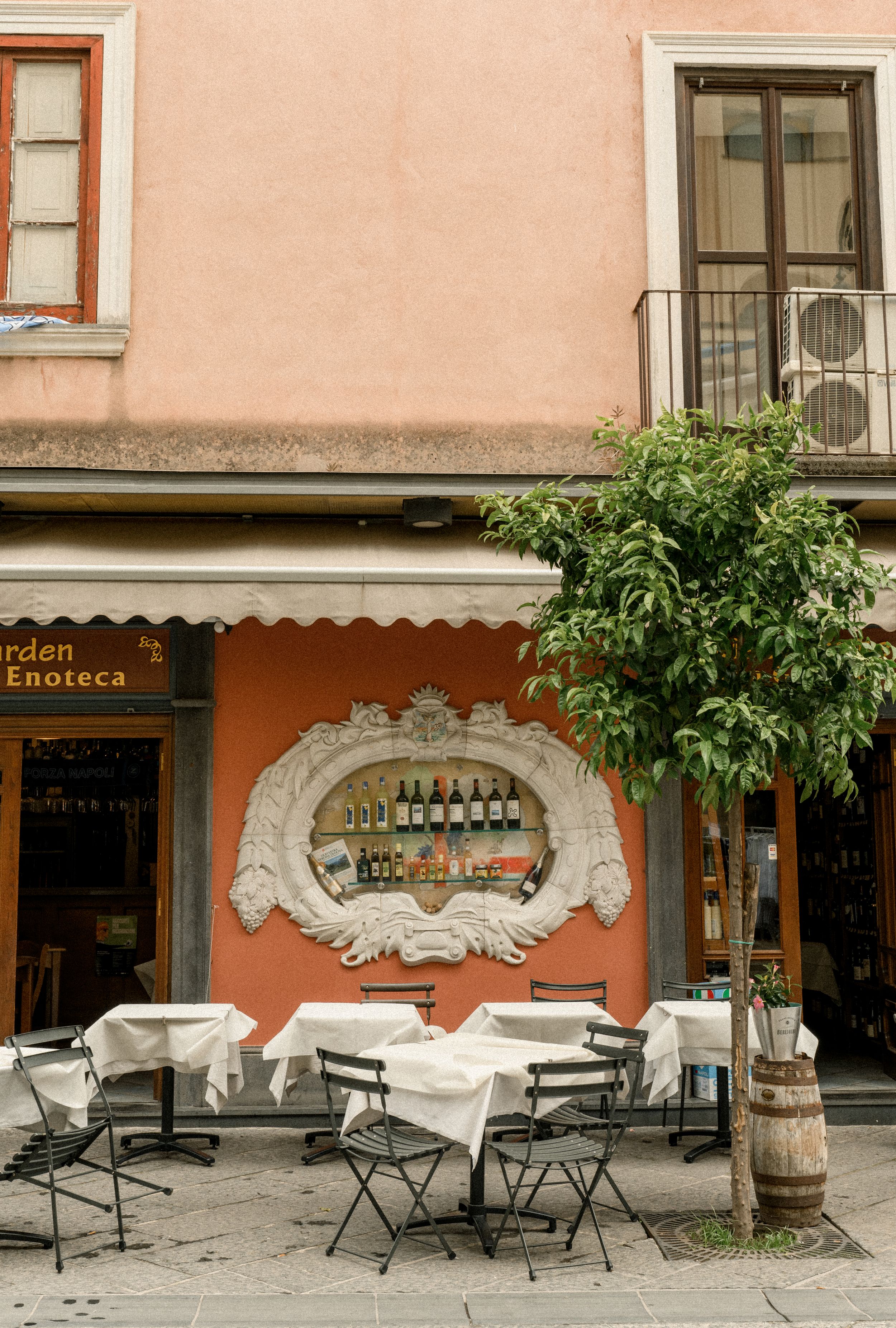 Outdoor terrace of an Italian enoteca with folding chairs and ornate wine display in a decorative wall niche.