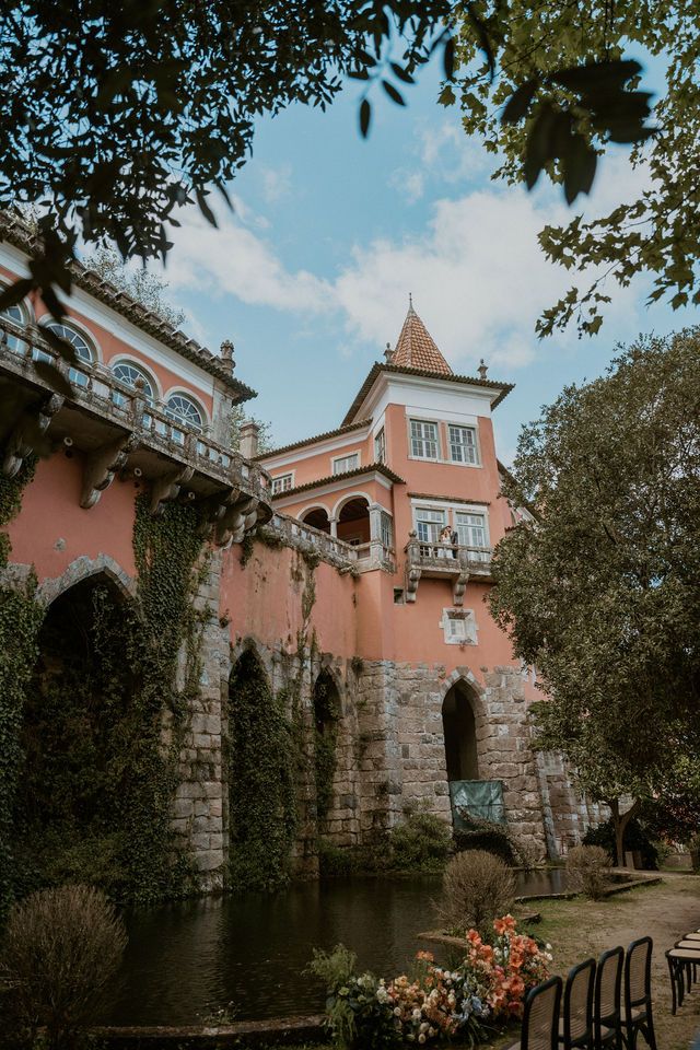 Romantic pink palace with stone foundation and turret reflecting in moat surrounded by lush gardens.