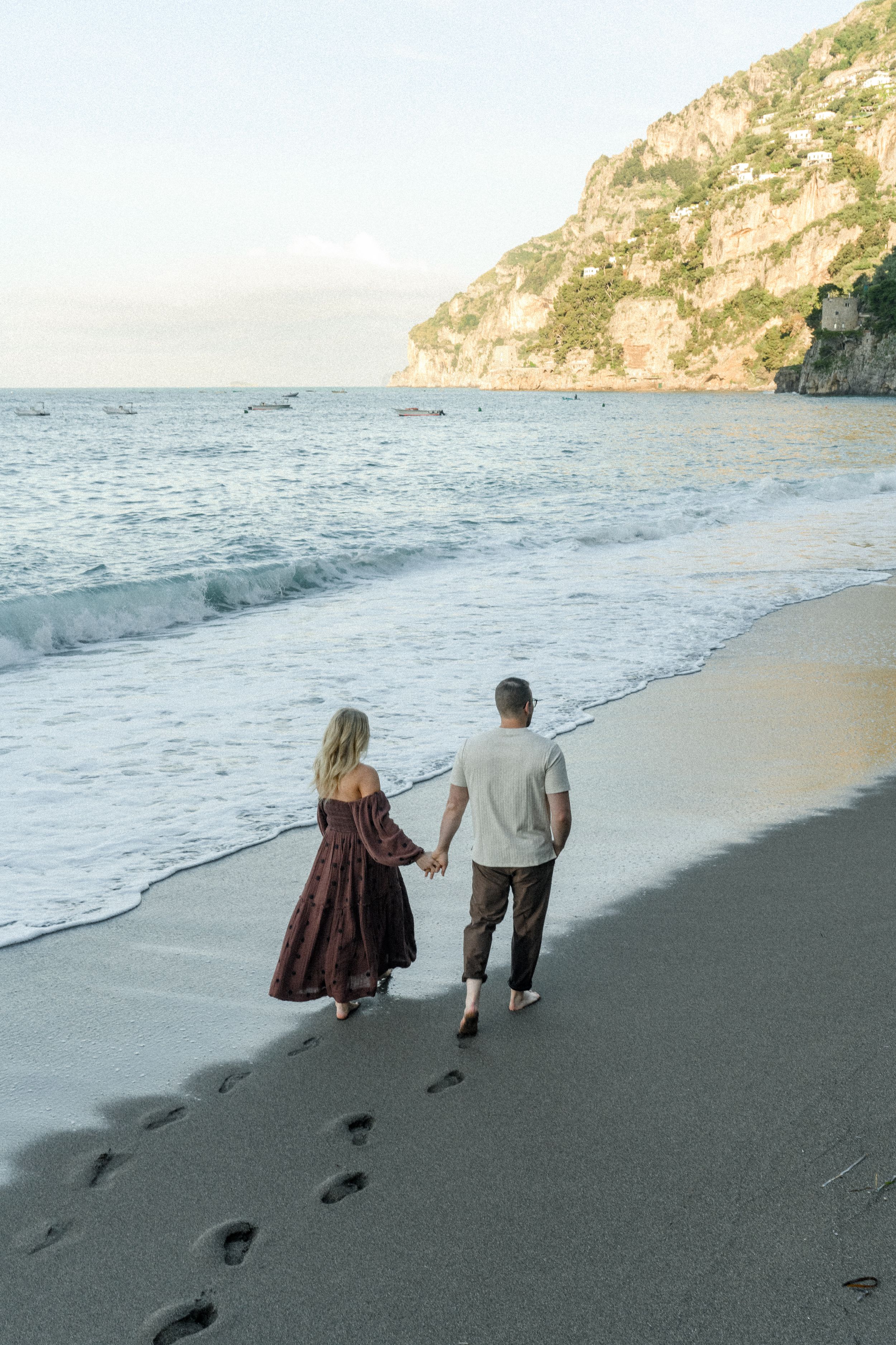 Couple walking hand in hand on beach leaving footprints in sand with cliffs in background.