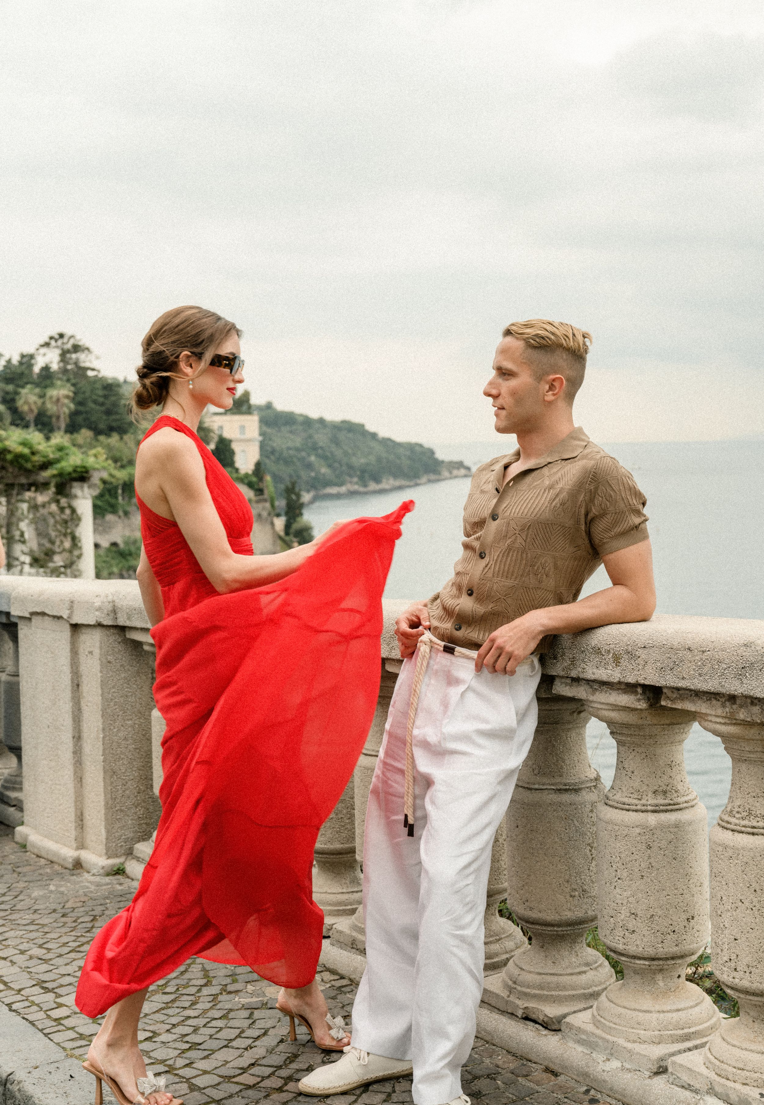 Red flowing dress catches wind as couple poses on coastal overlook with ocean view.