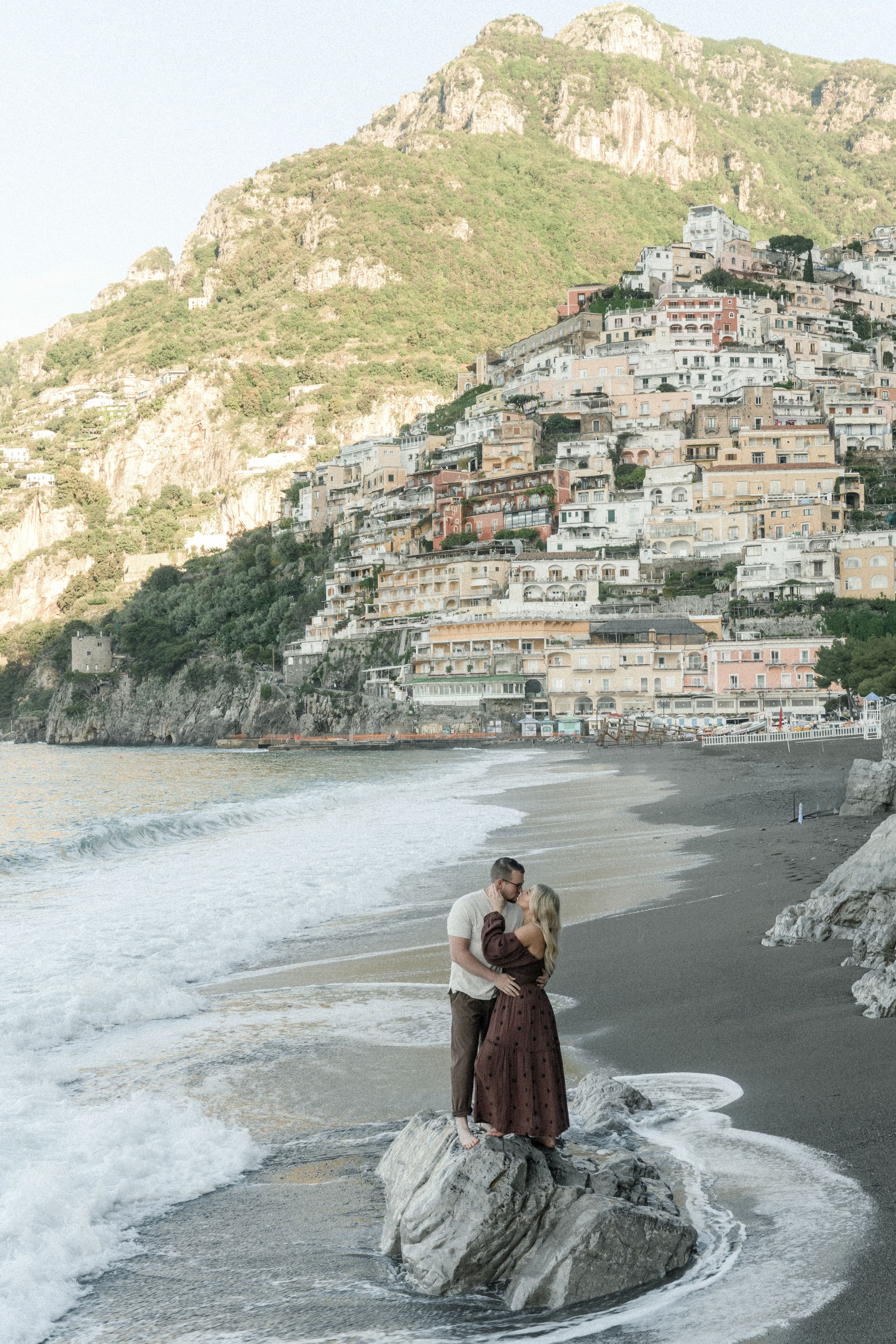 Couple standing on rocky beach with stunning view of hillside houses in Positano, Italy.