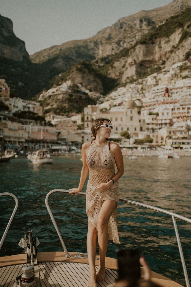 Woman in flowing dress on yacht deck with Positano coastal village backdrop in Italy.