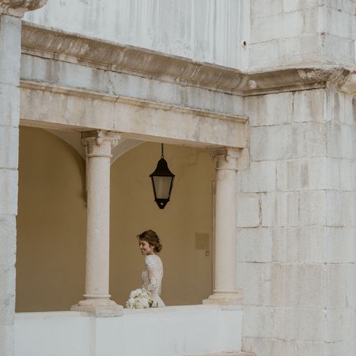 Bride with bouquet standing in stone archway with classical architecture details.