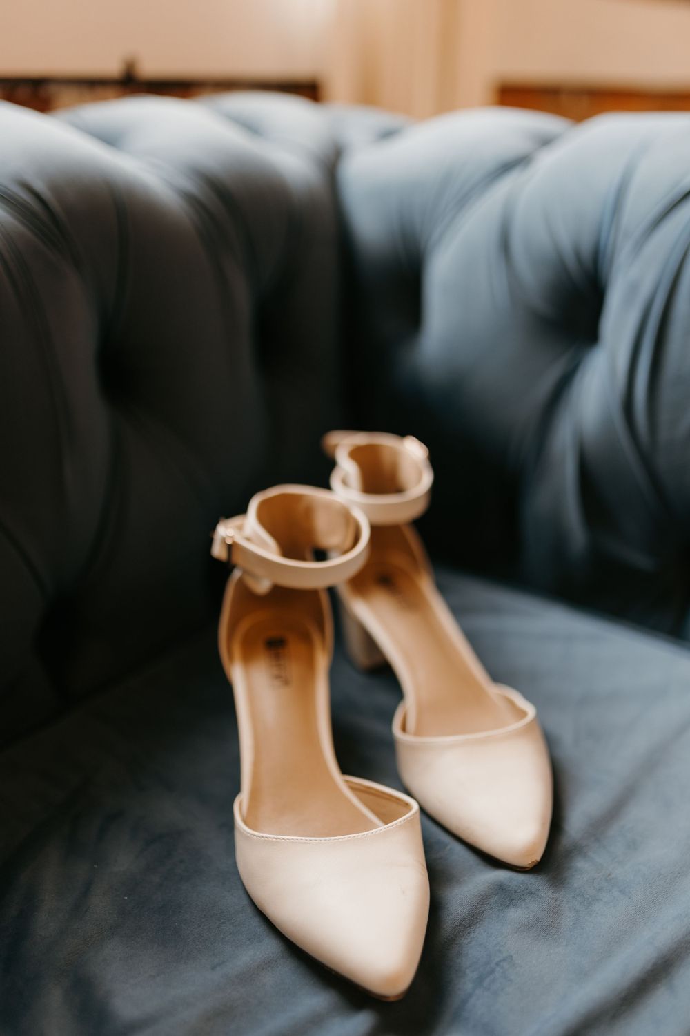 Elegant ivory pointed-toe bridal heels with ankle straps resting on a tufted blue velvet sofa.