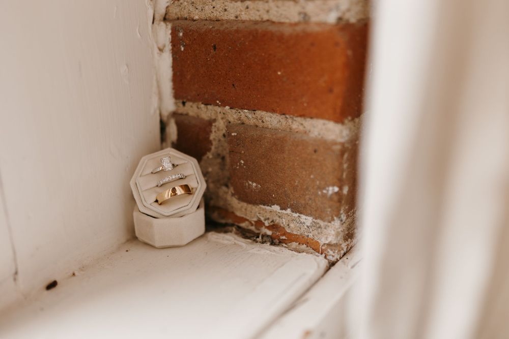 Gold wedding bands displayed in a white ring box nestled in a rustic brick corner windowsill.