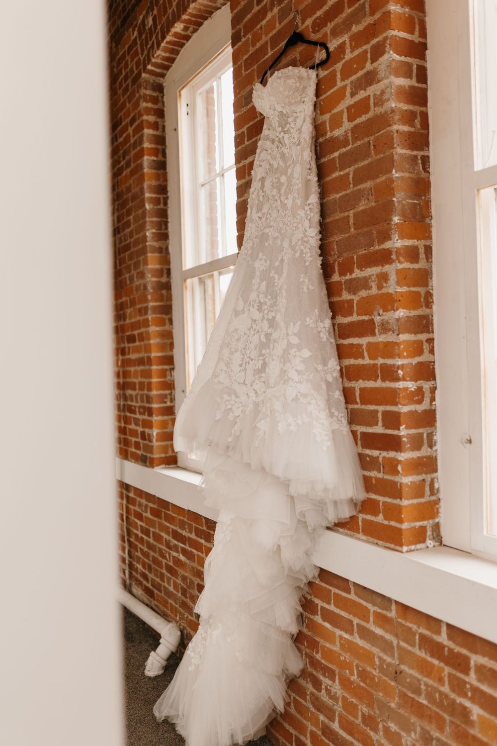 Strapless lace wedding gown with ruffled train hanging against a rustic exposed brick wall by a window.