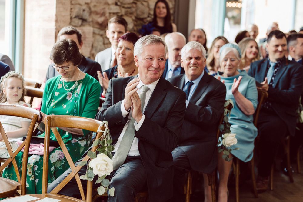 Wedding guests seated in wooden chairs watching ceremony in stone-walled venue.