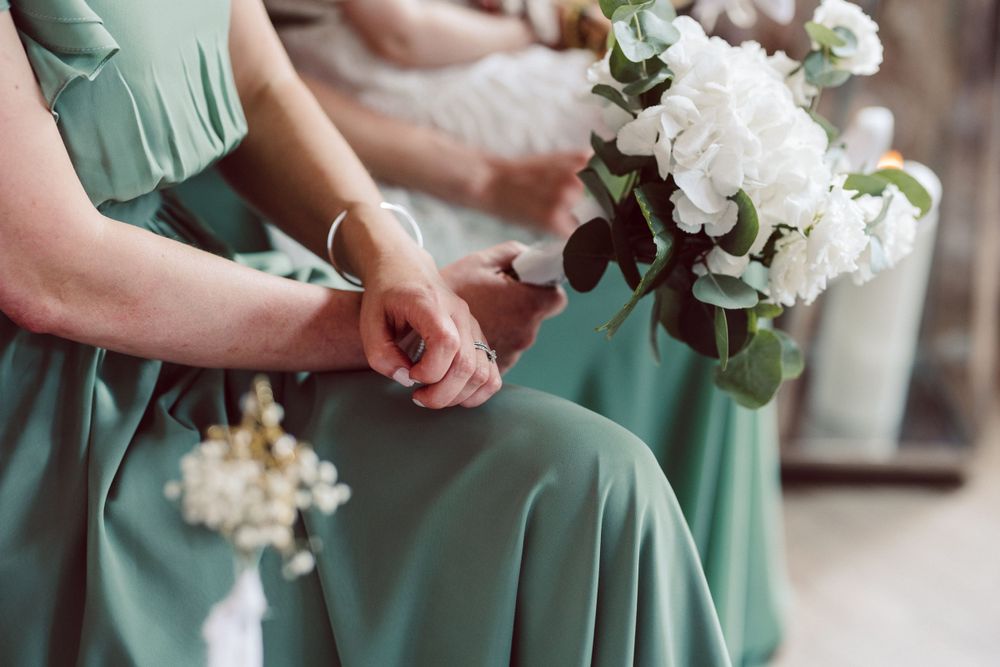 Bridesmaids in sage green dresses hold white floral bouquets at a wedding ceremony.