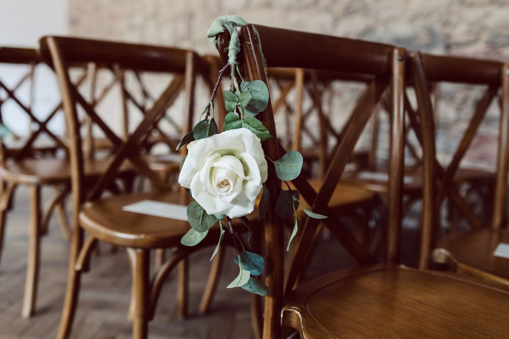 Wooden cross-back chairs decorated with white roses and greenery for a wedding ceremony.