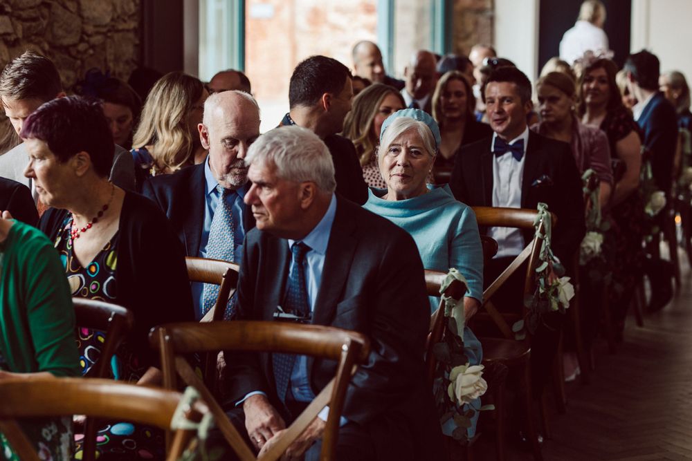 Wedding guests in formal attire seated at a rustic indoor ceremony venue.