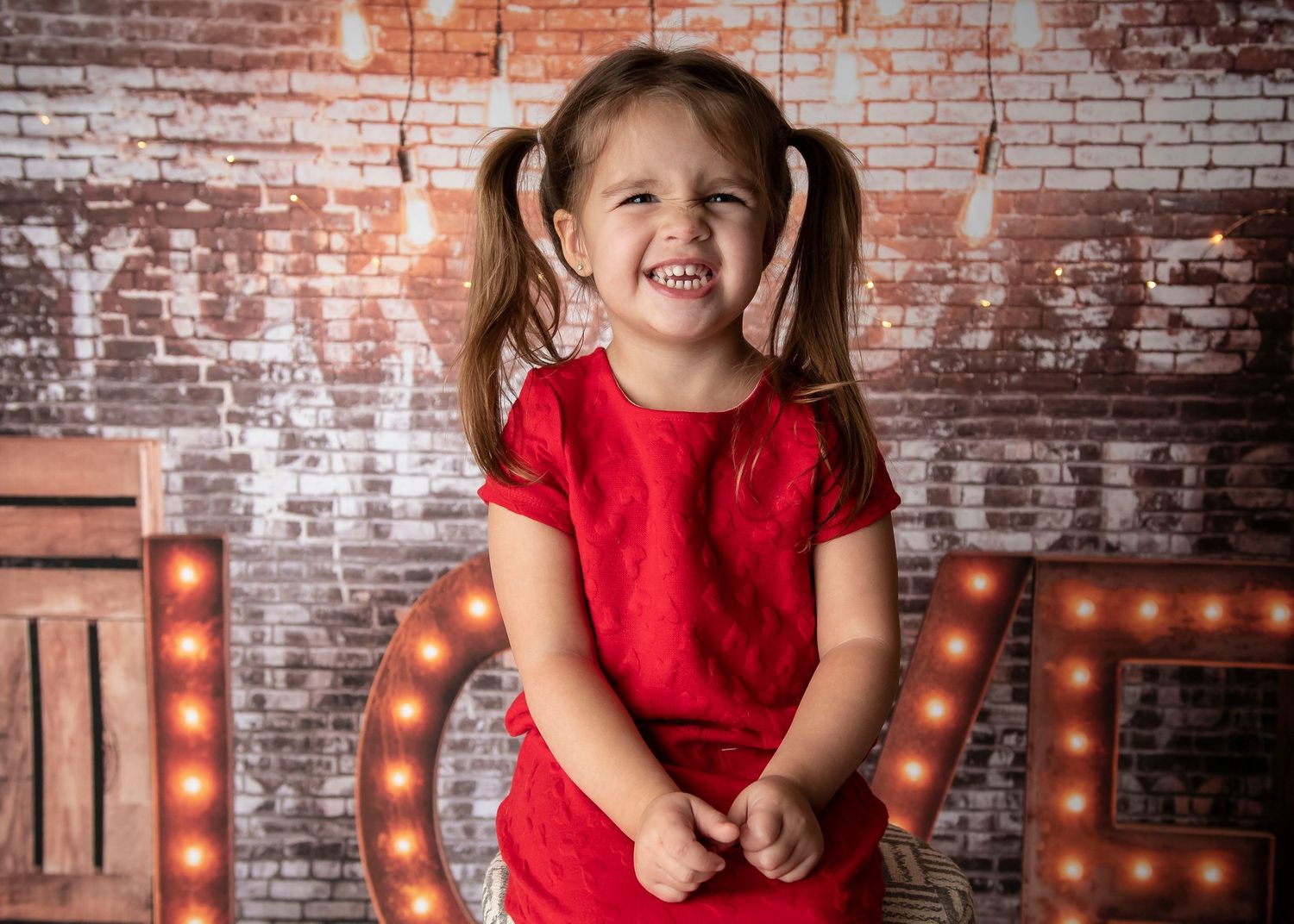 Child in red dress smiles joyfully against exposed brick wall with illuminated LOVE letters.