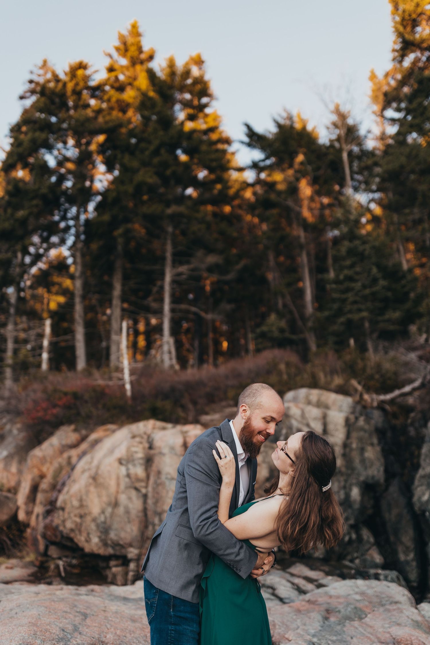 Couple embracing on rocky outcrop with autumn forest backdrop at sunset.