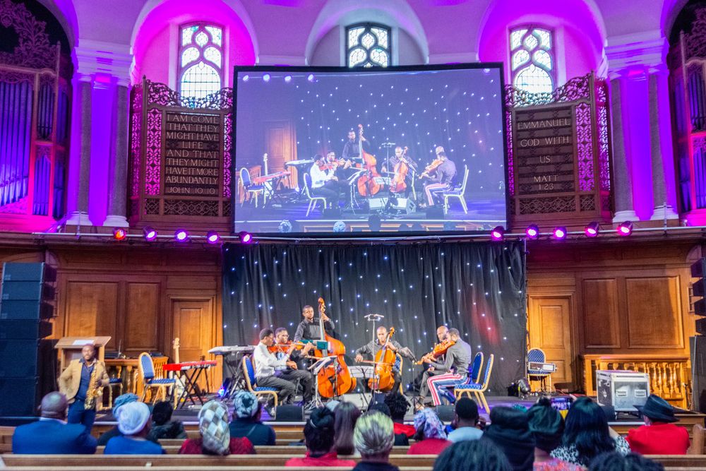 String ensemble performing on stage in grand church hall with large projection screen.