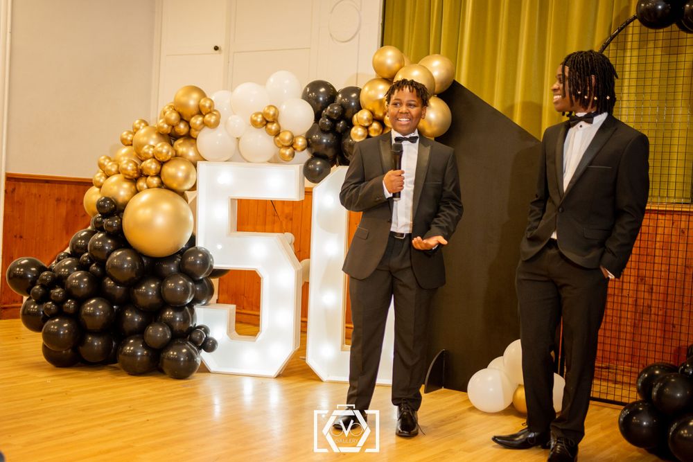Two men in suits speak on stage at a 50th birthday party celebration.