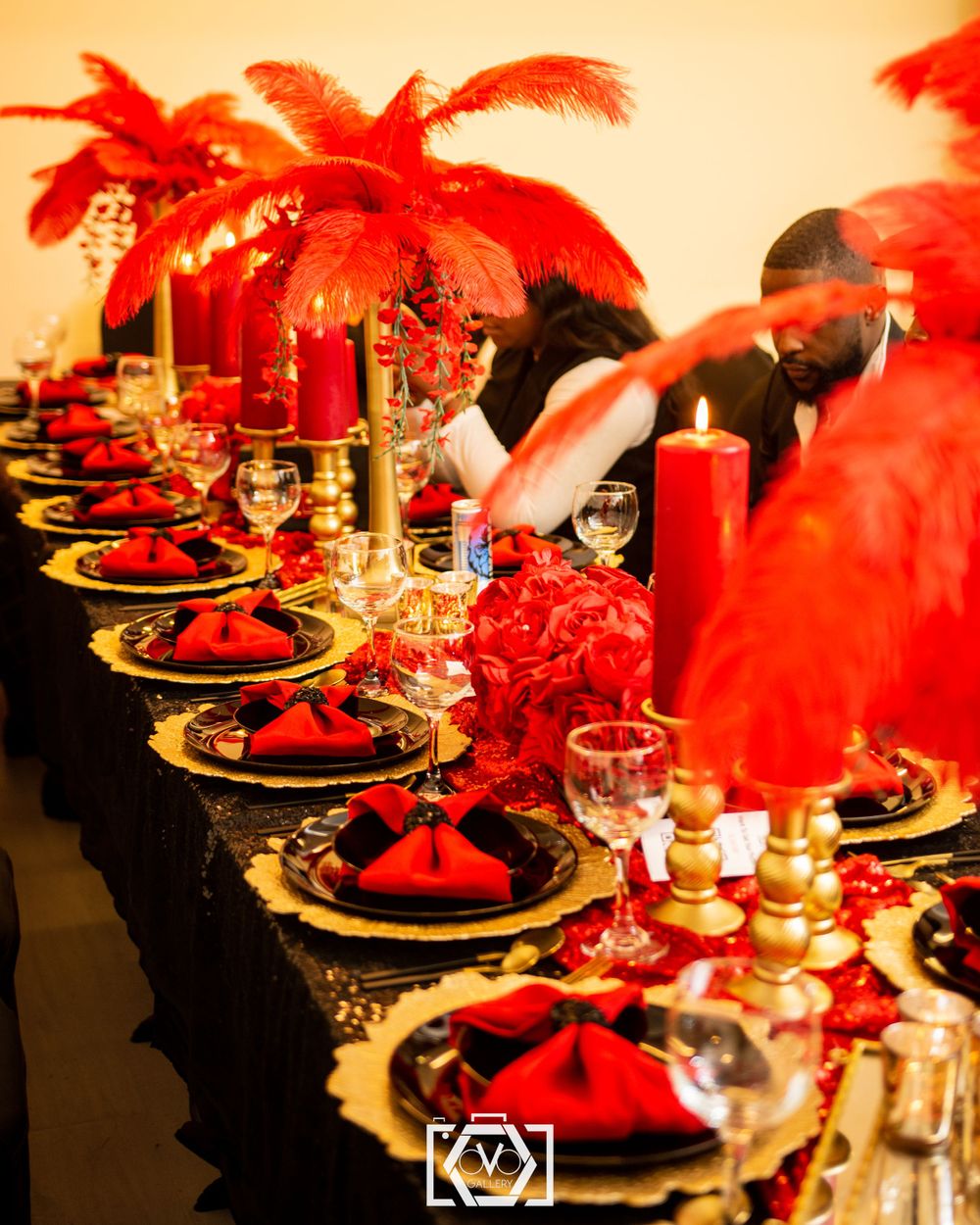 Long banquet table with red feather centerpieces, gold candle holders, red napkins, and black sequin runner.