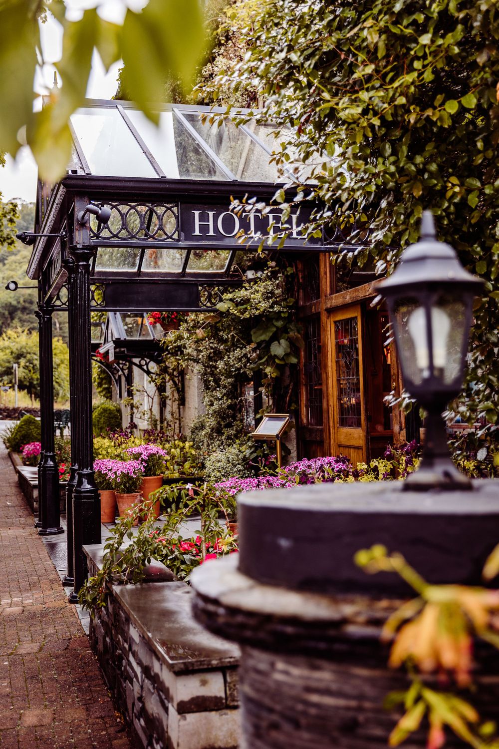 Quaint hotel entrance with ornate signage and flowering plants along a brick pathway.