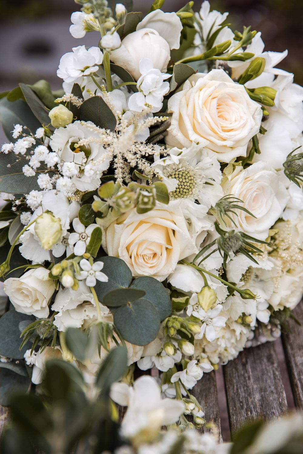 Elegant white wedding bouquet with cream roses, baby's breath, and eucalyptus leaves arranged on rustic wood.
