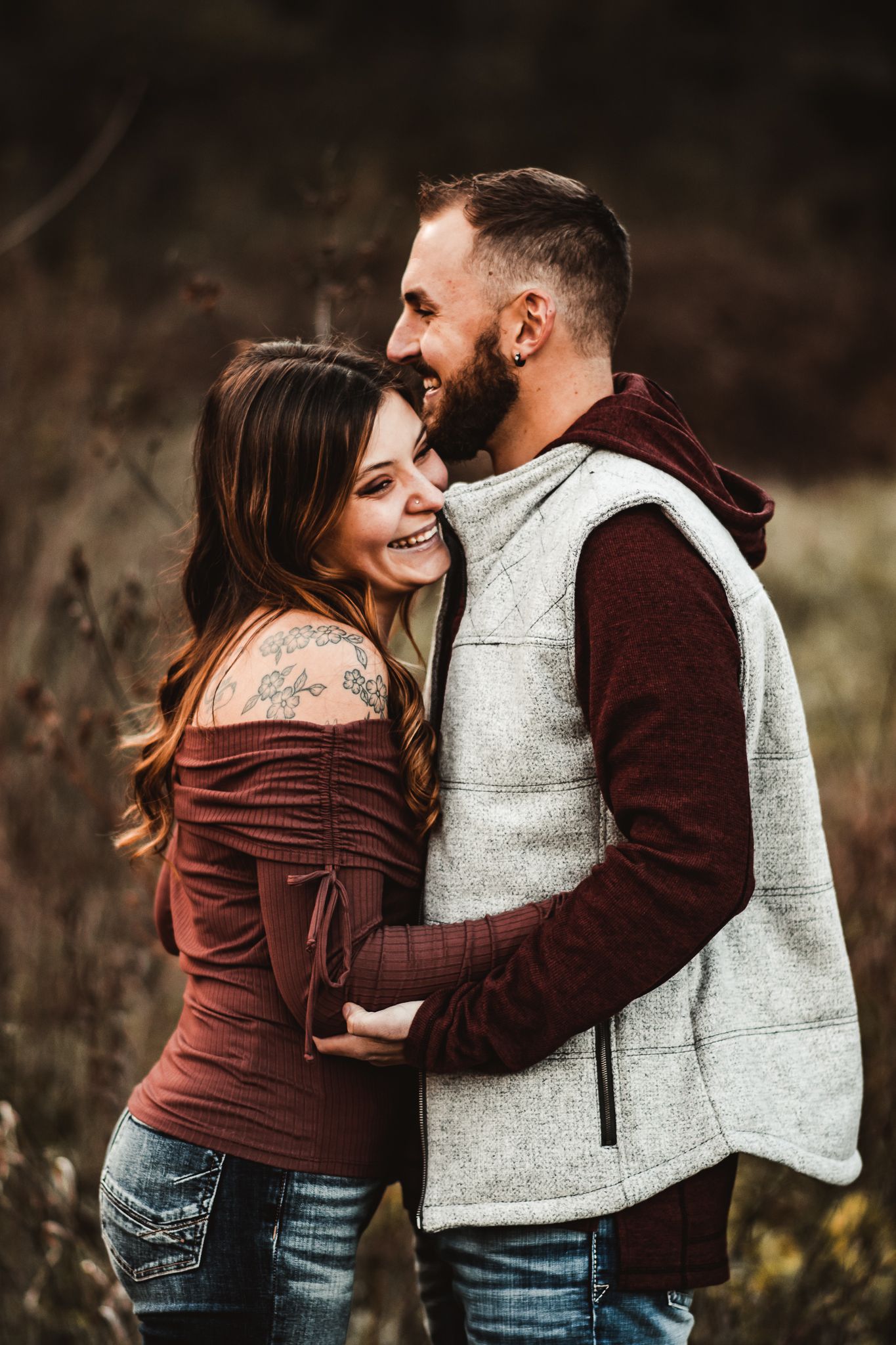 Romantic couple embracing and laughing together in casual fall attire against natural backdrop.