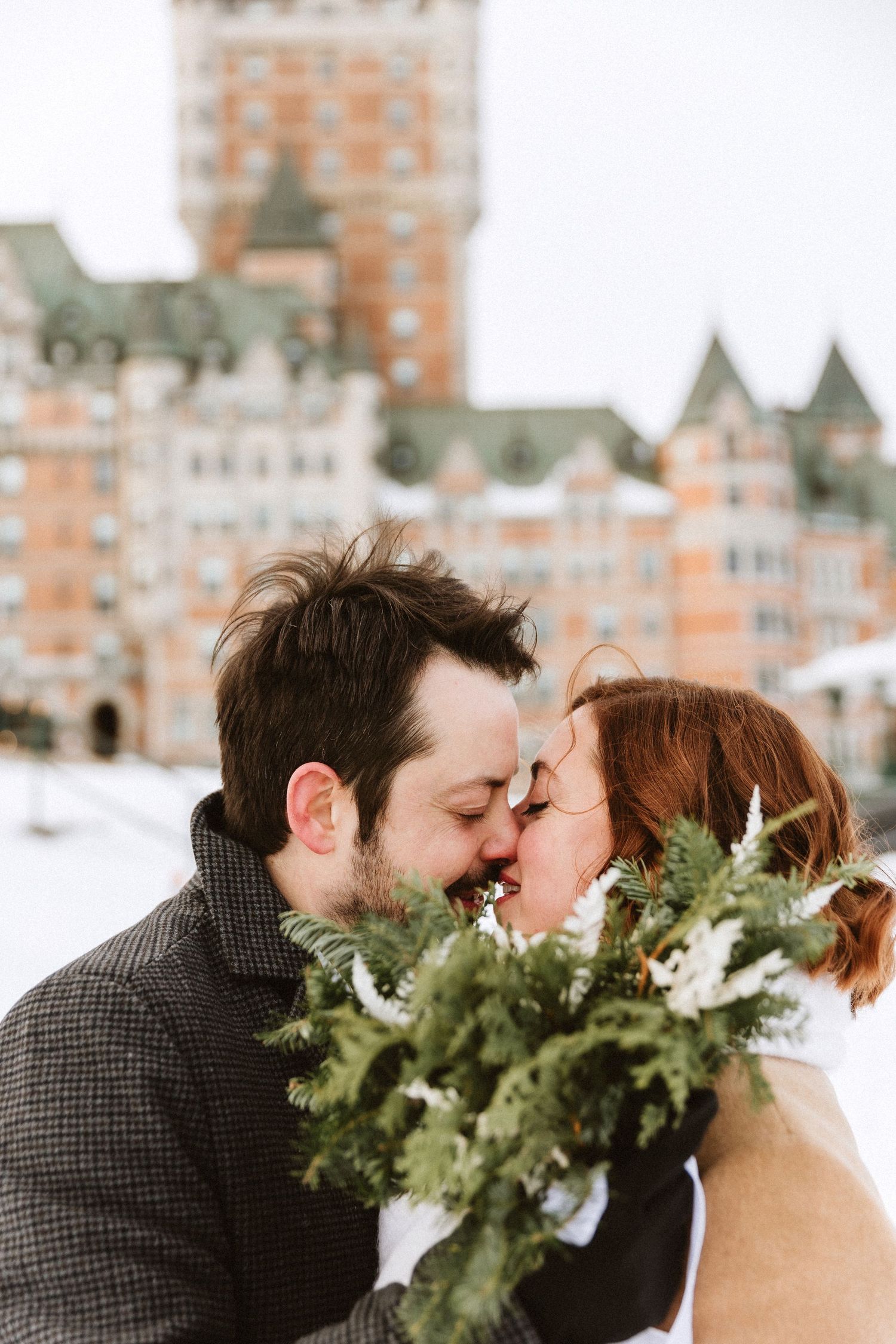 A romantic winter wedding couple shares a kiss with evergreen bouquet against a snowy chateau backdrop.