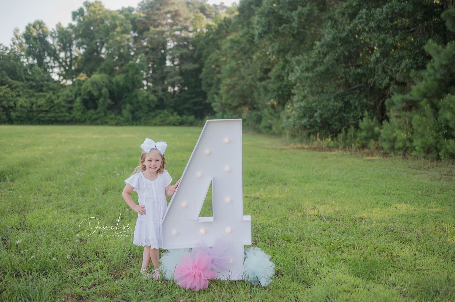 Small child in white dress standing next to large number 4 decoration outdoors.