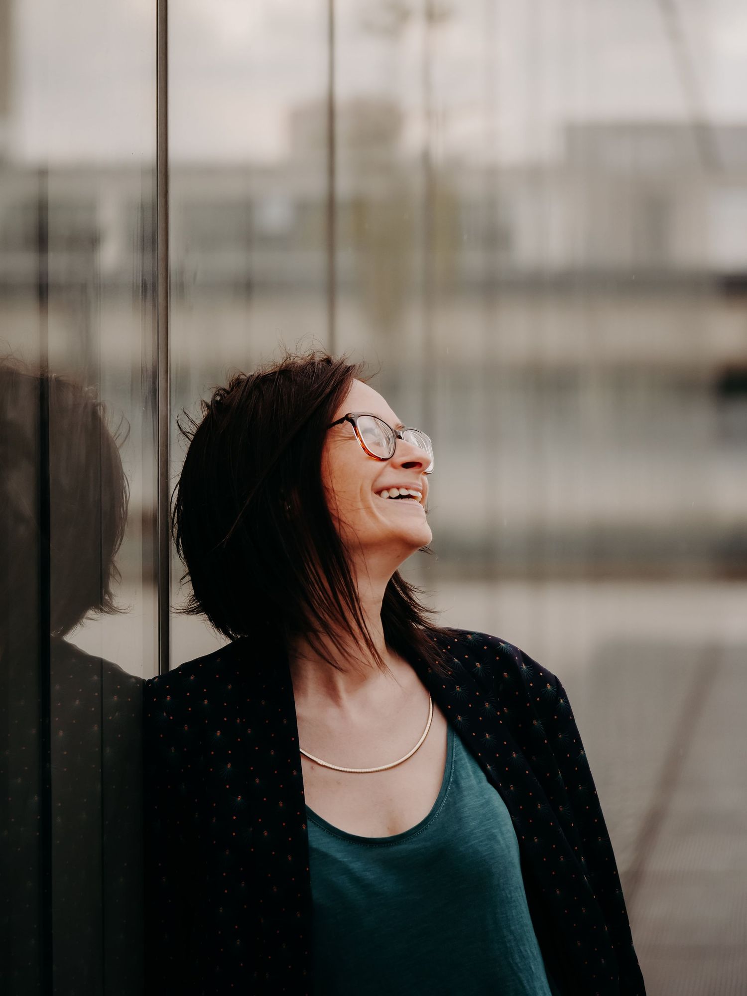 A woman wearing glasses and a black cardigan laughs joyfully against a blurred background.