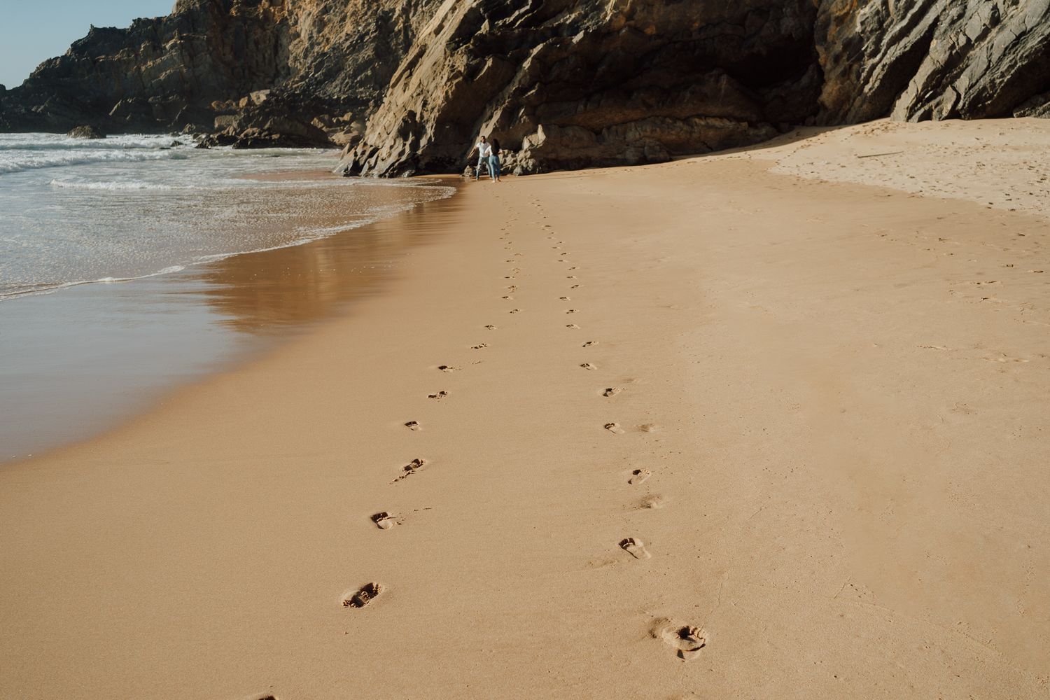 Footprints trail along a sandy beach beside rocky cliffs and ocean waves.