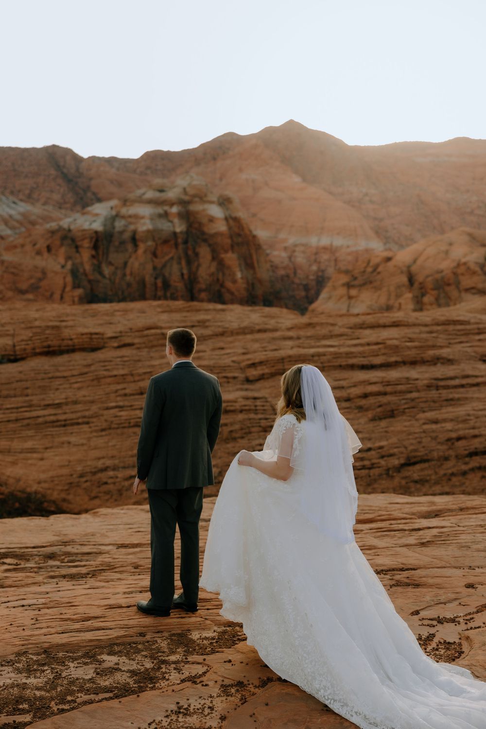 Bride in flowing white gown and groom overlook majestic desert canyon vista.