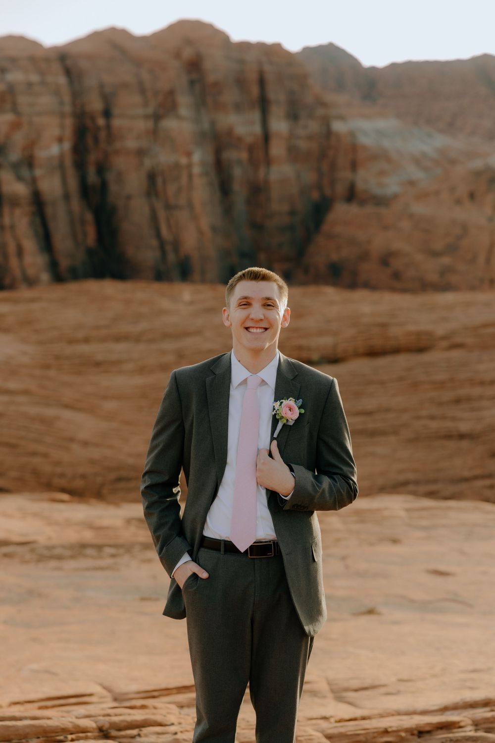 A groom in formal attire with pink tie stands against red rock cliffs in the desert.