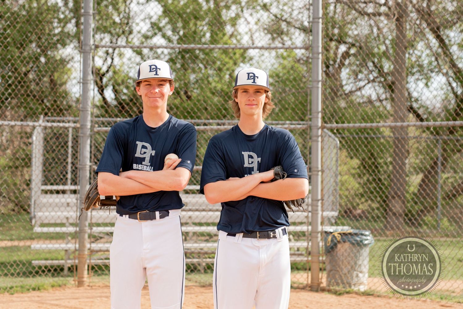 Two baseball players in navy uniforms stand with crossed arms near a chain link fence on a sunny day.
