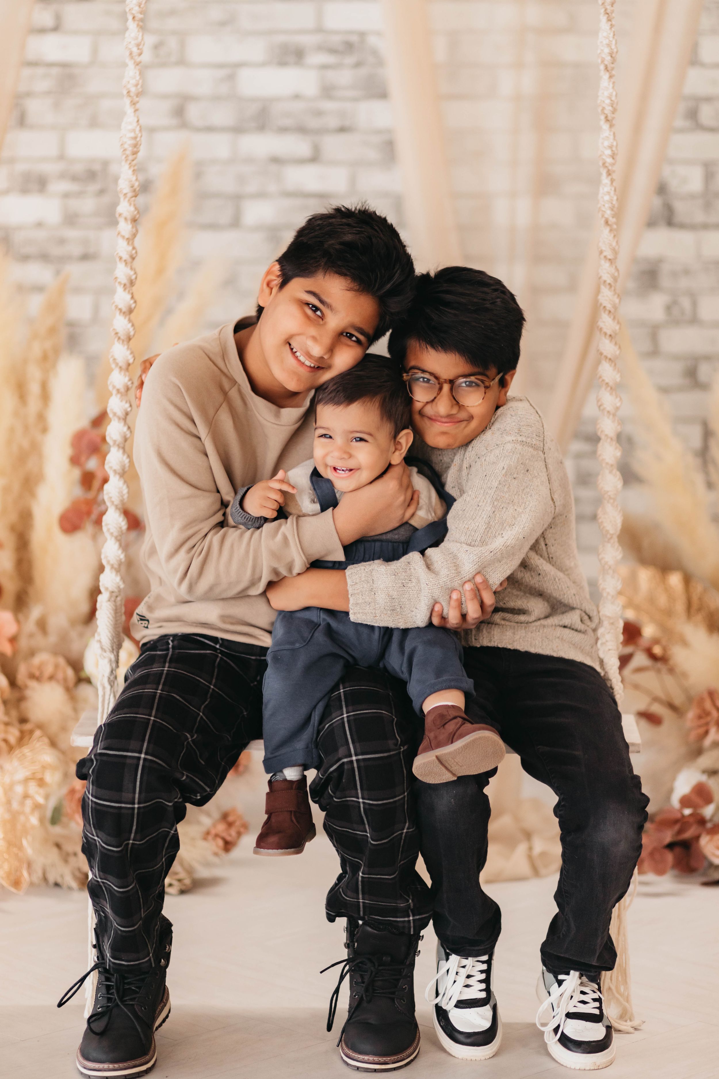 Three siblings share a sweet moment together on a rope swing against a neutral brick wall backdrop.